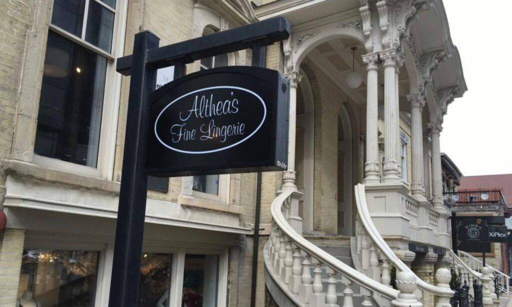 The image shows the exterior of Althea's Fine Lingerie, located at 777 N. Jefferson Street in Milwaukee. The building has a historic architectural style with light-colored brick walls and ornate columns. A grand, curved staircase with intricate railings leads up to the entrance. The black store sign with white lettering reads 'Althea's Fine Lingerie' and hangs from a post attached to the building. A glimpse of the storefront windows reveals part of the interior display, hinting at a cozy and elegant shopping experience. The overall scene is inviting and showcases the boutique's unique, upscale setting.